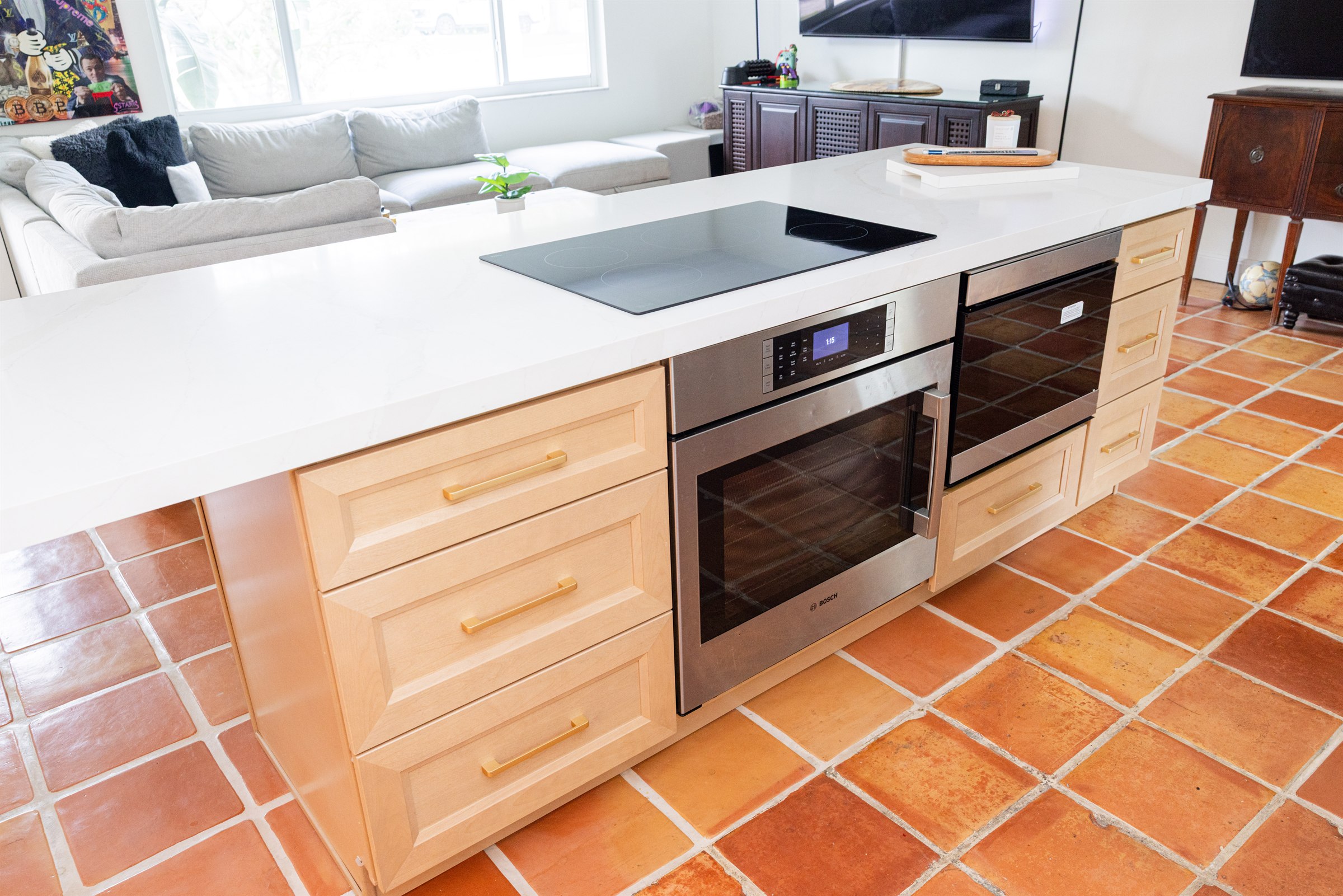 Kitchen island with double wall ovens, induction cooktop, and wide terracotta-tile walkway to the living room