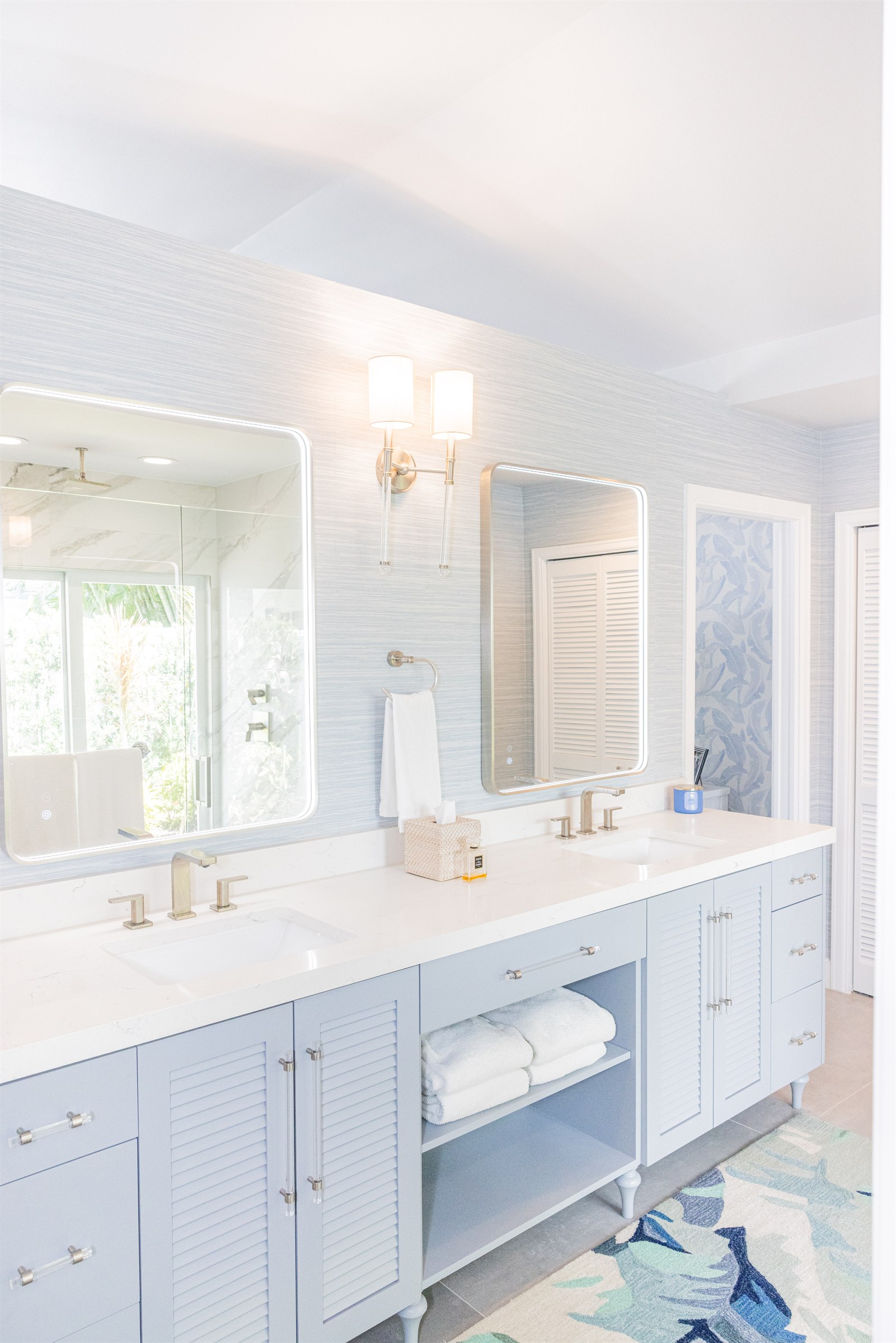 Blue louvered vanity with grasscloth wallpaper and a mirror reflecting the shower