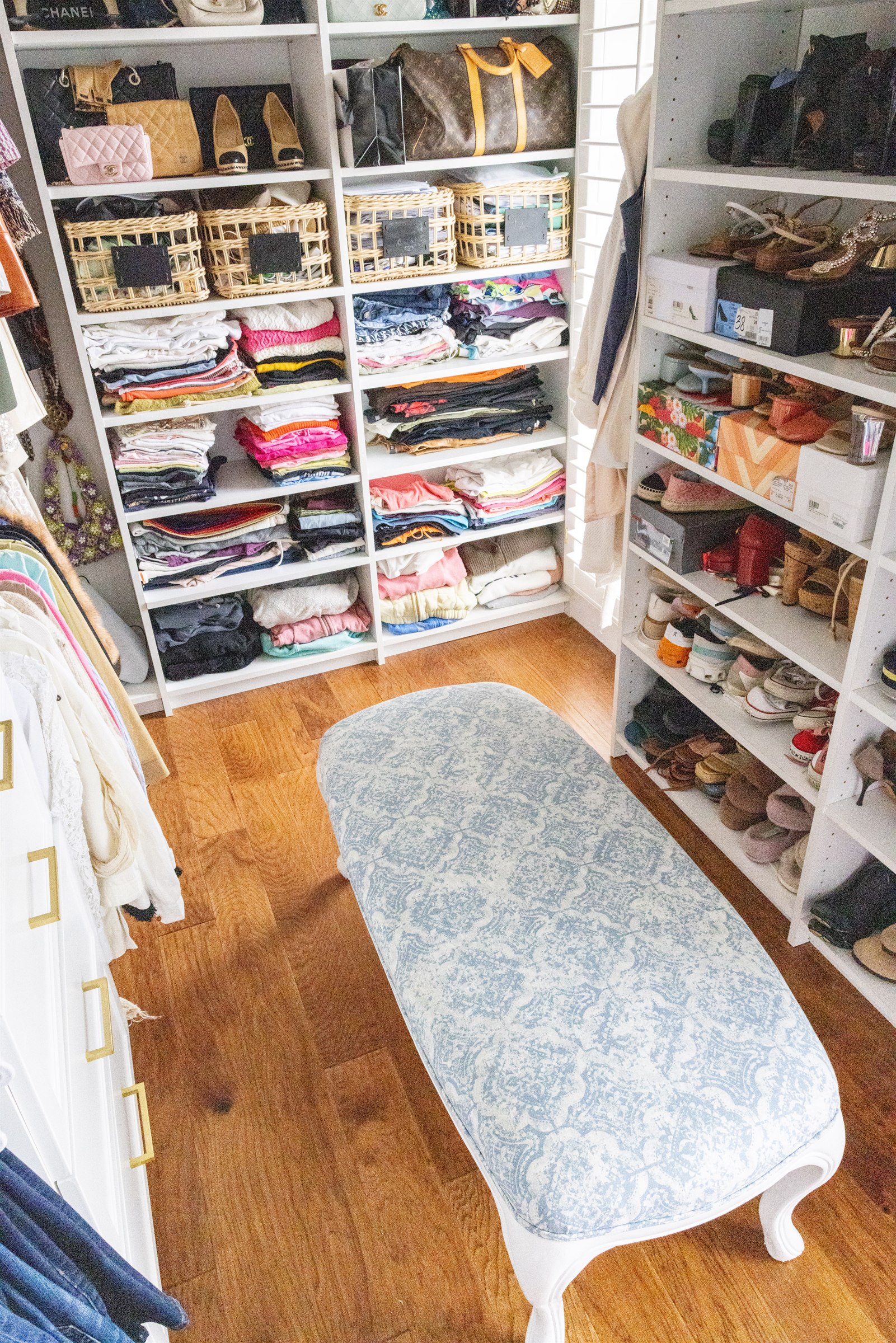 Walk-in closet with folded clothes shelving, handbag display, and blue upholstered bench