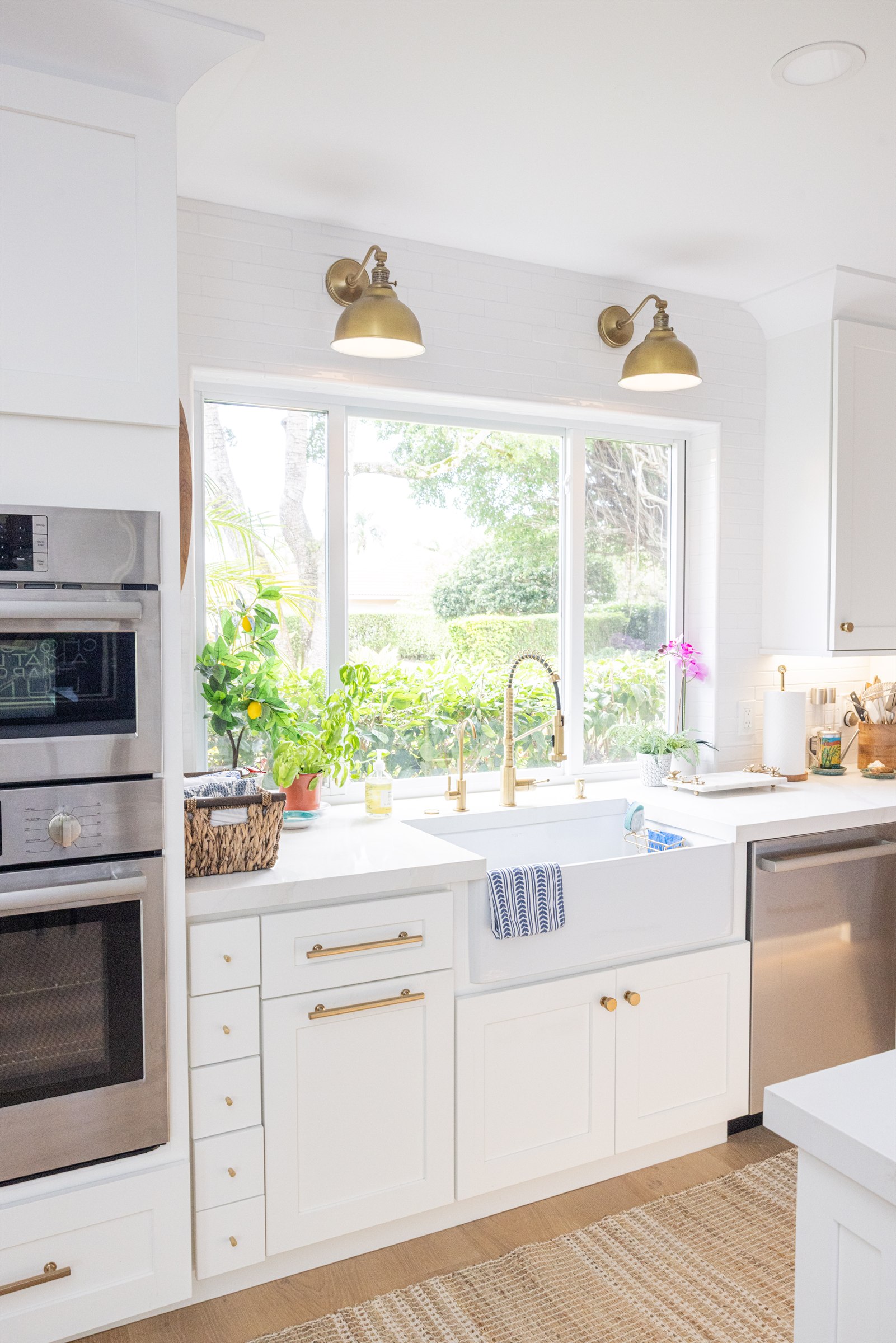 White farmhouse sink beneath a garden window flanked by brass sconces
