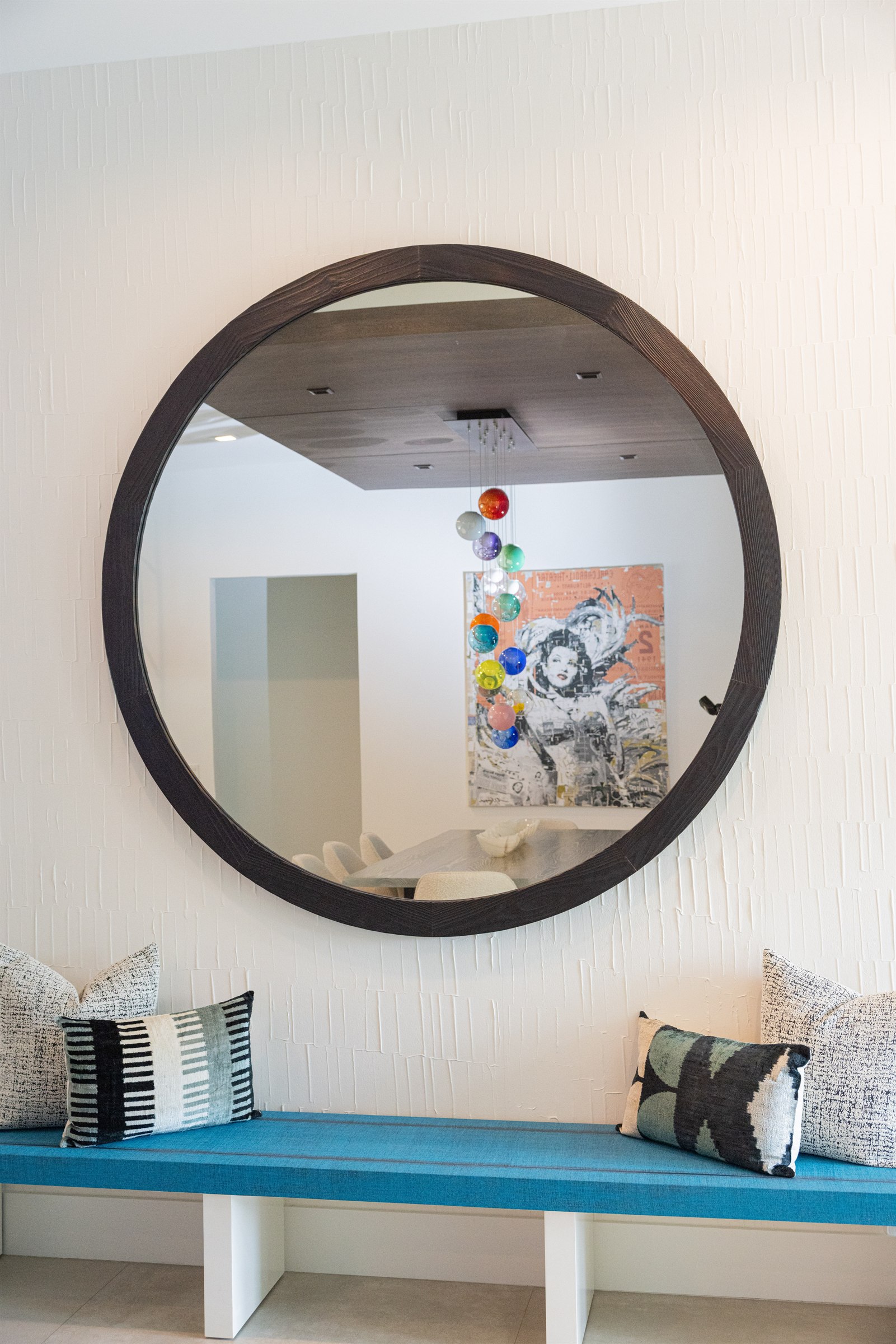 Round mirror above a blue bench seat reflecting the dining room and colorful artwork