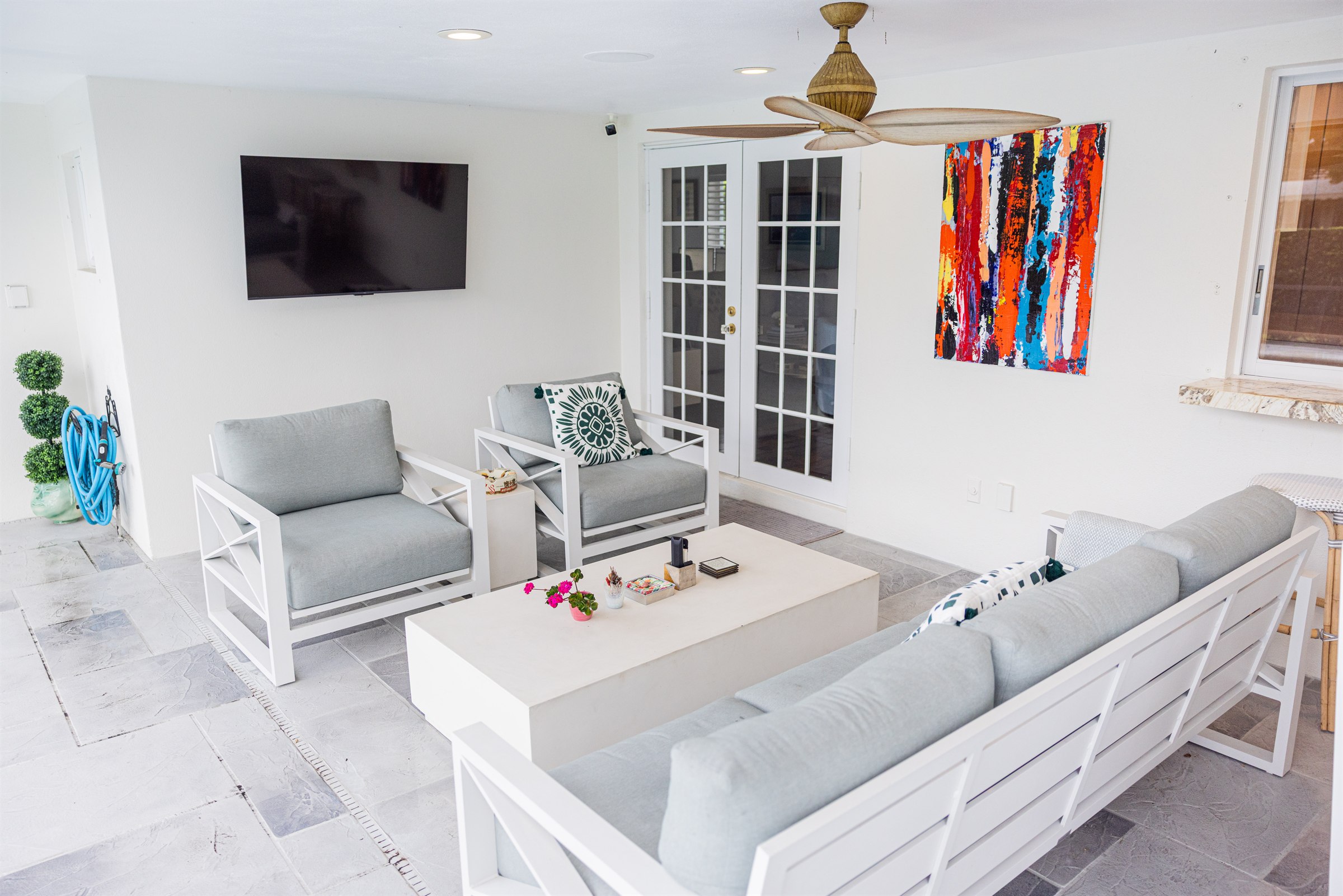 Outdoor living room with gray cushioned lounge chairs, sofa, wall-mounted TV, and colorful abstract artwork