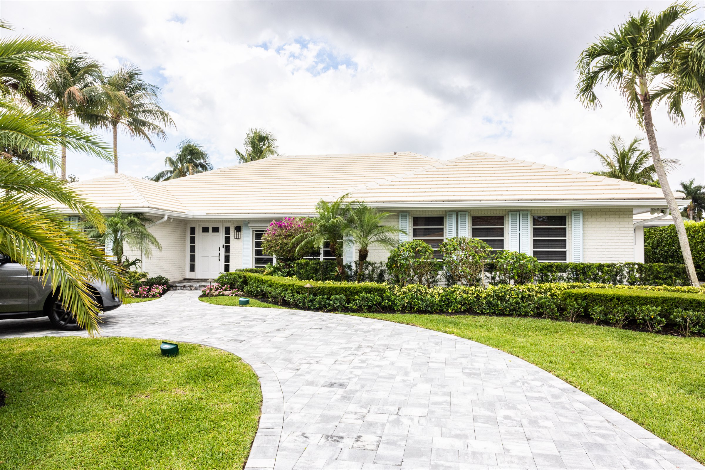 White coastal home exterior with pale blue shutters, curving paver driveway, and tropical landscaping