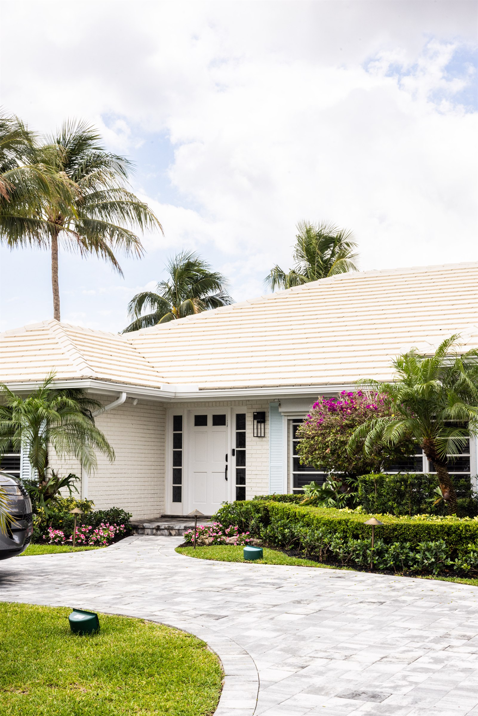 Front entrance detail with paneled white door, light blue shutter, and paver walkway framed by palms and bougainvillea